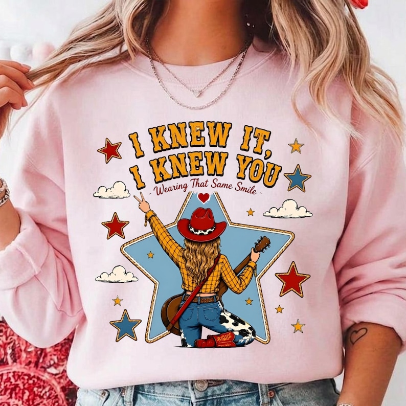 May include: A light pink crewneck sweatshirt featuring a graphic of a person wearing a red cowboy hat, yellow plaid shirt, and cow-print trousers, holding a guitar. The text reads I KNEW IT, I KNEW YOU Wearing That Same Smile, surrounded by stars.