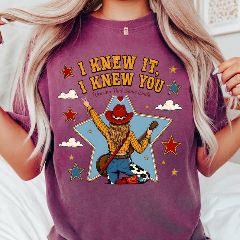 May include: A purple t-shirt featuring a graphic of a person in a red cowboy hat, yellow plaid shirt, and cow-print jeans holding a guitar. The text reads I KNEW IT, I KNEW YOU, Wearing That Same Smile, surrounded by stars and clouds.