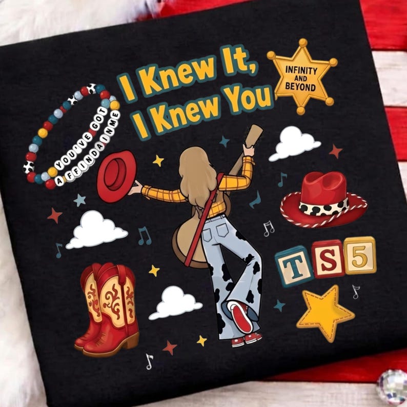 May include: A black t-shirt featuring a graphic of a person with long hair holding a guitar, wearing a yellow plaid shirt and cow-print pants. The text reads I KNEW IT, I KNEW YOU, INFINITY AND BEYOND, and TS5. Includes a beaded bracelet, cowboy boots, and hats.