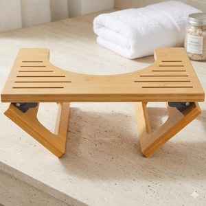 May include: A light brown bamboo toilet stool featuring a U-shaped cutout in the centre and horizontal slats on the top surface. The stool has two foldable legs with dark metal hinges, providing a stable base for use in a bathroom setting.