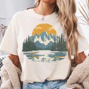May include: A cream-colored short-sleeve t-shirt featuring a graphic print of a mountain range, a forest of pine trees, and a lake under a large yellow sun. The design has a distressed texture and is centered on the front of the garment.