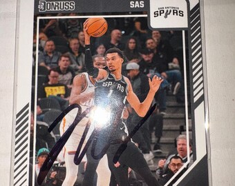 Może przedstawiać: Karta kolekcjonerska do koszykówki Donruss przedstawiająca Victora Wembanyamę z San Antonio Spurs w akcji. Karta pokazuje zawodnika trzymającego piłkę do koszykówki, autograf czarnym tuszem na środku oraz napis Victor Wembanyama.