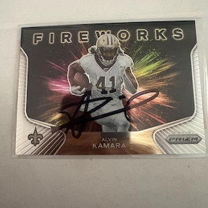 Peut inclure: Une carte de sport Panini Prizm Fireworks représentant Alvin Kamara portant un maillot blanc avec le numéro 41. La carte présente un fond noir avec des rayons lumineux colorés, une signature en encre noire et le logo des New Orleans Saints.