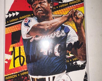 Può includere: Una carta da baseball con Hank Aaron in uniforme dei Braves, che tiene una mazza. La carta ha uno sfondo colorato con il testo "HANK AARON" e "AARON". La carta è racchiusa in una custodia protettiva.