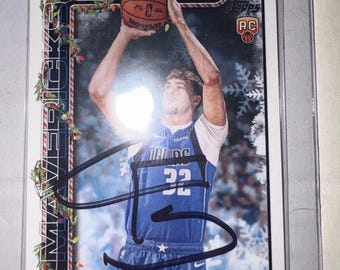 Może przedstawiać: Karta kolekcjonerska do koszykówki Topps przedstawiająca Coopera Flagga w niebieskiej koszulce Dallas Mavericks z numerem 32, trzymającego piłkę nad głową. Karta ma świąteczną ramkę z girlandami i płatkami śniegu oraz czarny autograf.