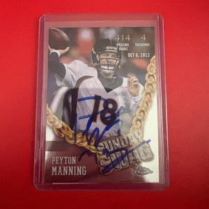 May include: A Peyton Manning football card in a protective case. The card features Manning in a Denver Broncos uniform, with the number 18. The card includes the text "414 Passing Yards" and "4 Touchdowns" from Oct 6, 2013, and a signature.