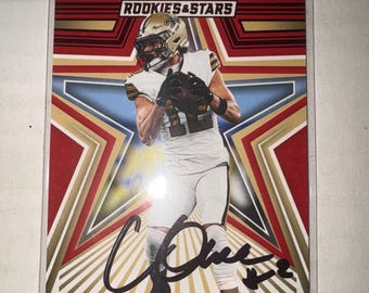 May include: A Rookies & Stars trading card featuring Chris Olave of the New Orleans Saints. The card displays a player in a white uniform with a gold helmet, set against a red, white, and blue star background. It includes a black ink autograph.