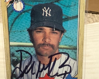 May include: A Topps baseball card featuring Don Mattingly wearing a dark blue New York Yankees cap. The card has a wood-grain border, a blue background, and a black ink autograph across the center. Text includes Don Mattingly, American League, and All Star.