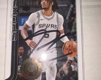 Może przedstawiać: Karta kolekcjonerska Topps przedstawiająca Stephona Castle'a w białej koszulce San Antonio Spurs, trzymającego piłkę do koszykówki Wilson. Karta zawiera autograf wykonany czarnym tuszem oraz złoty emblemat NBA All-Rookie Team.