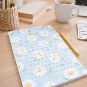 May include: A light blue notepad with a daisy floral pattern and the word "Notes" in gold. A gold pen rests on the notepad. The notepad is on a wooden desk with a cup of coffee, a keyboard, and a woven pen holder.