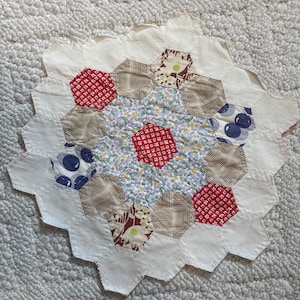 May include: A white fabric quilt with a hexagonal pattern. The quilt is made of various fabrics with different patterns, including red, blue, and brown. The quilt is hand-stitched and has a vintage look.