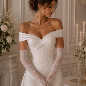 Könnte beinhalten: Weißes schulterfreies Brautkleid mit herzförmigem Ausschnitt und eng anliegendem Oberteil. Das Kleid hat zarte Spitzenhandschuhe und einen fließenden Rock. Die Braut trägt eine Halskette mit einem kleinen Anhänger.