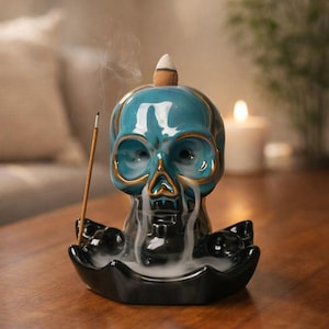 May include: A teal ceramic skull incense burner with gold accents. The skull-shaped burner has a cone incense on top and a stick of incense in the front. Smoke is emanating from the skull's mouth and eyes, and the burner sits on a black dish.