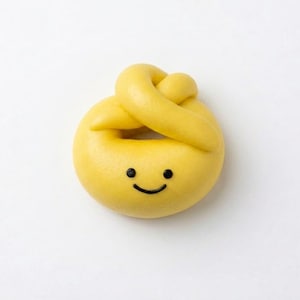 May include: A bright yellow, knotted dough-like object with a smooth, rounded surface. It features a simple, hand-drawn black ink face consisting of two small circular eyes and a curved line representing a smiling mouth centered on the front.