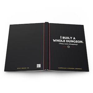May include: A black hardcover campaign and session journal with white text reading I BUILT A WHOLE DUNGEON. (They went shopping.) and a small die icon. The back cover features the text EPIC BOON CO. and a small square QR code.