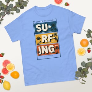 May include: Light blue t-shirt with a graphic design that says "SURFING" in three stacked blocks. The design includes palm trees and the text "LIFE OF THE WIND" and "OCEAN IS CALLING".