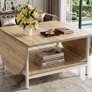 May include: A light wood-tone coffee table with a white metal frame. The table has a lower shelf with books and a tray with a cup and saucer. A vase of yellow flowers sits on the table. The table has drop-leaf sides.