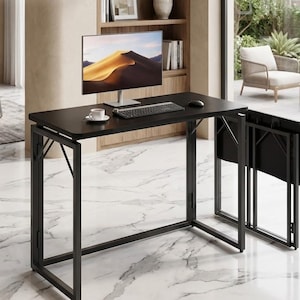 May include: A black folding desk with a rectangular tabletop and a metal frame. A computer monitor, keyboard, and mouse sit on the desk. A white teacup and saucer are also on the desk. The desk is set up in a well-lit room.