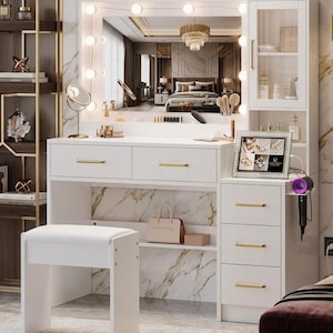 May include: A white vanity with a large mirror framed by lights. The vanity has drawers, a shelf, and a stool. A small cabinet is on the right side. The mirror reflects a bedroom with a chandelier.