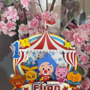 May include: A multi-layered paper cake topper featuring a circus tent design with a blue glitter number one, the name Elian in blue and white, and colorful cartoon characters including a blue-haired clown, a pink cat, and a brown bear.