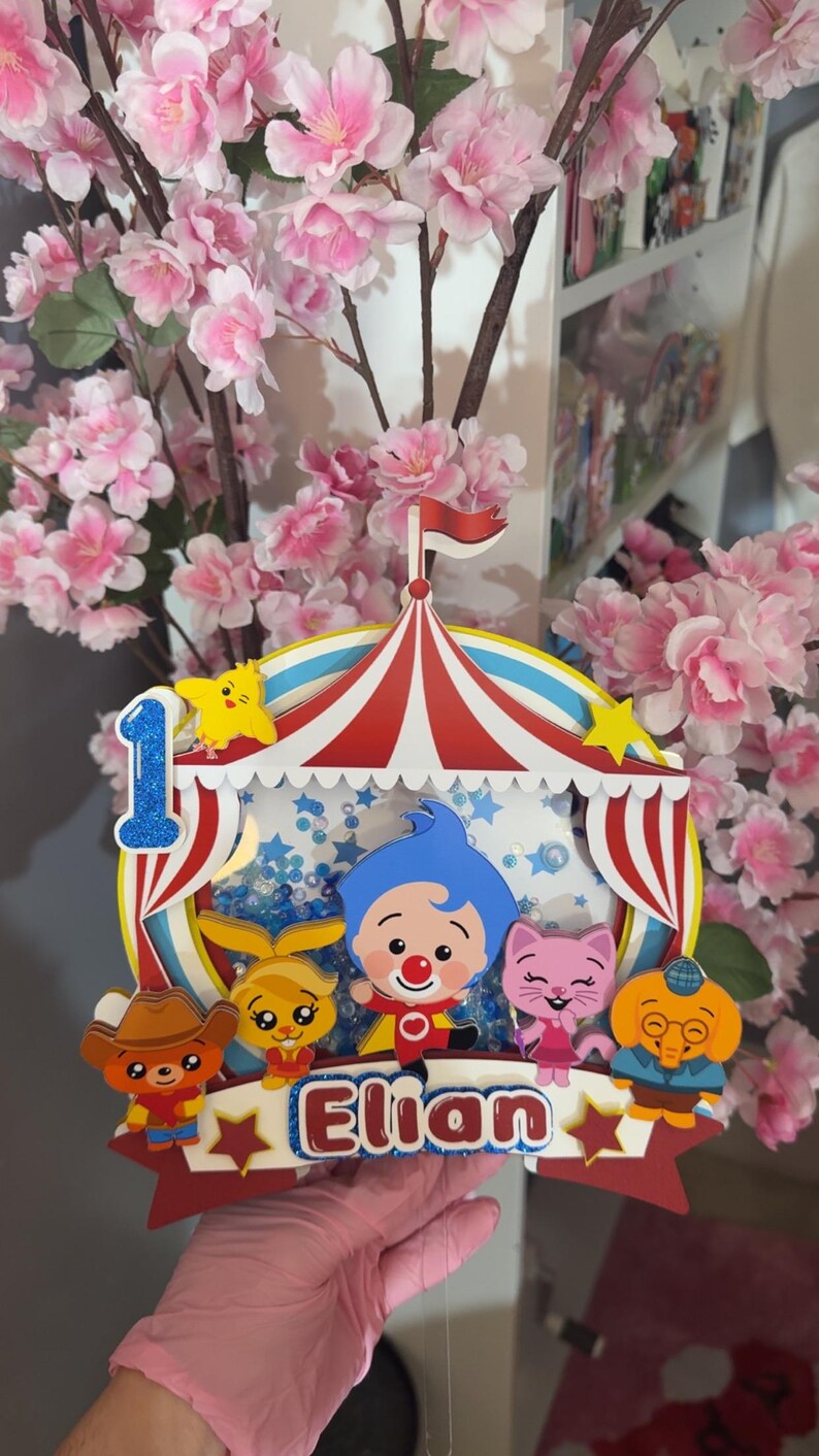 May include: A multi-layered paper cake topper featuring a circus tent design with a blue glitter number one, the name Elian in blue and white, and colorful cartoon characters including a blue-haired clown, a pink cat, and a brown bear.
