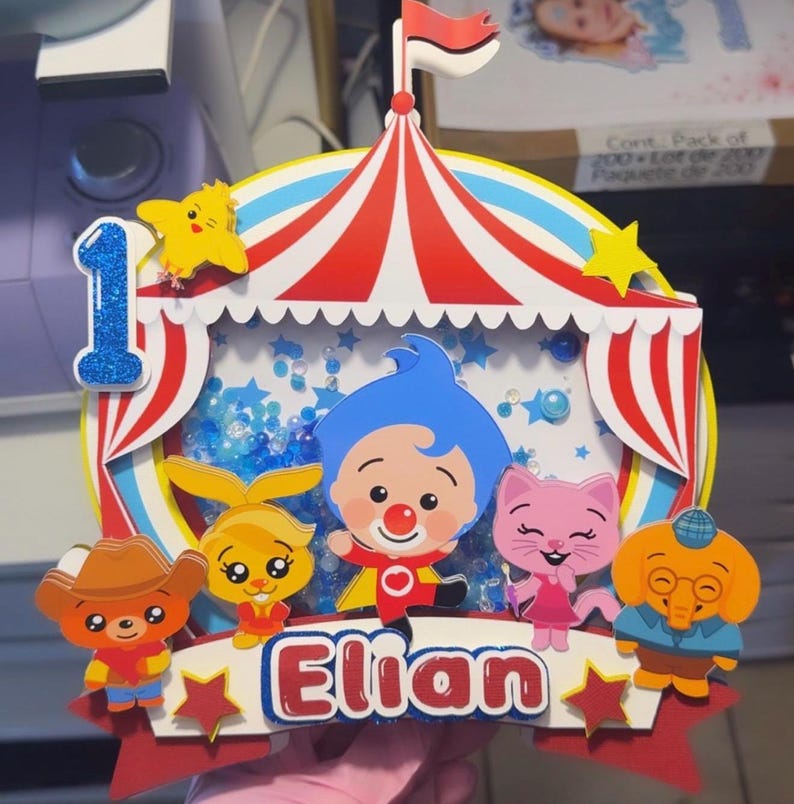 May include: A layered paper cake topper featuring a circus tent design with red and white stripes, a blue glittery number one, and a shaker element filled with blue sequins. The name Elian is printed in red, surrounded by cartoon characters.