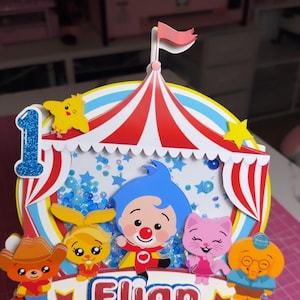 May include: A multi-layered paper cake topper featuring a circus tent design with red and white stripes. It includes a blue glittery number one, a central shaker element with blue sequins, and cartoon characters. The name Elian is printed in red.