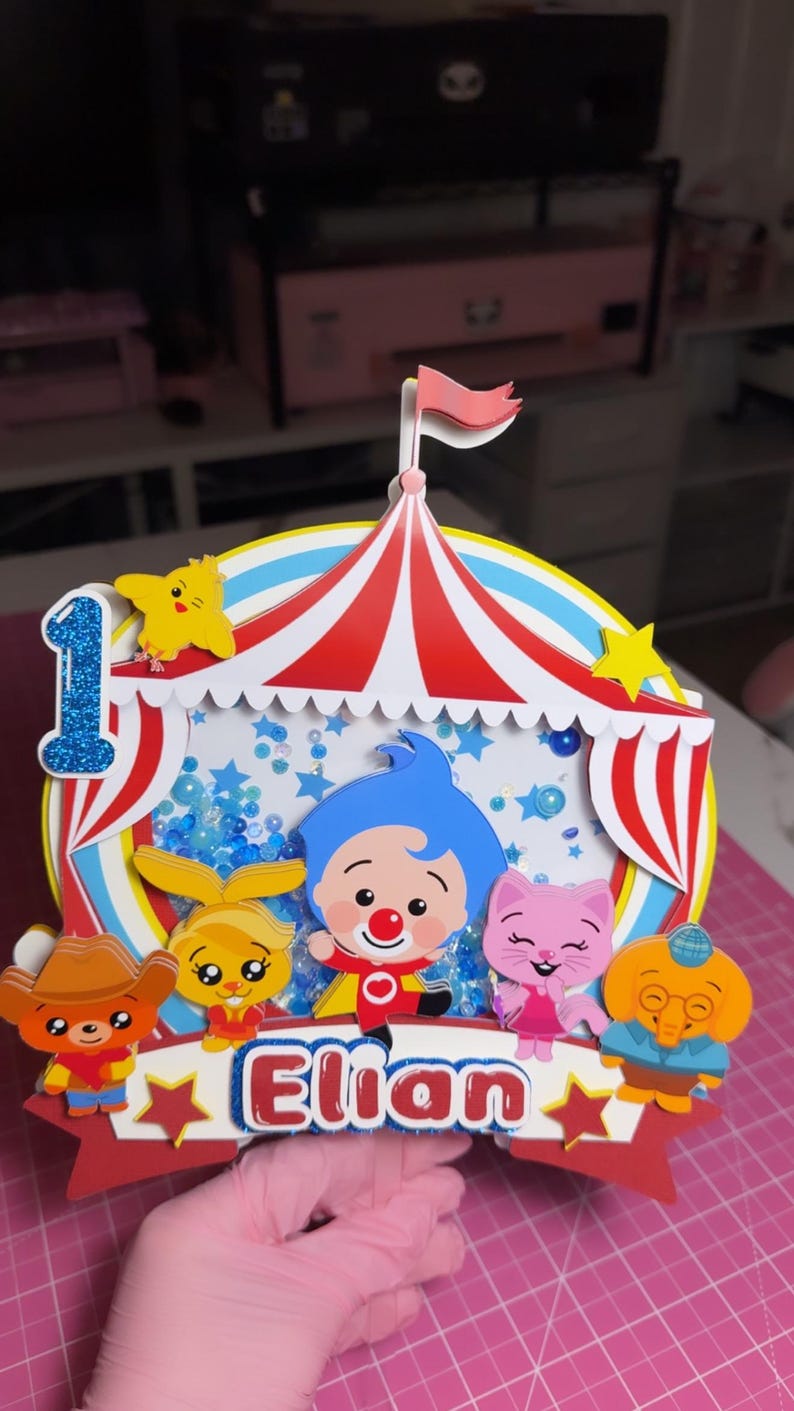 May include: A multi-layered paper cake topper featuring a circus tent design with red and white stripes. It includes a blue glittery number one, a central shaker element with blue sequins, and cartoon characters. The name Elian is printed in red.