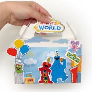 May include: A white cardboard box decorated with Sesame Street characters, including Elmo, Cookie Monster, and Oscar the Grouch. The box has a handle and features the text "Jovan Jr's WORLD" and "Sesame Street". Balloons and a number 1 are also visible.