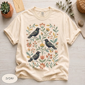 May include: An ivory short-sleeve t-shirt featuring a central graphic of three black birds perched among stylized green vines, orange flowers, and red berries. The shirt has a crew neckline and a Comfort Colors brand label at the inner collar.