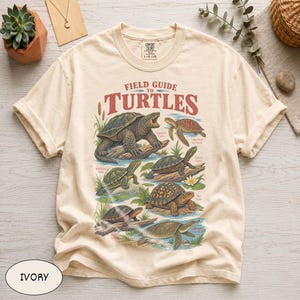 May include: An ivory short-sleeve t-shirt featuring a graphic print titled Field Guide to Turtles. The design displays six detailed illustrations of various turtle species in natural settings with text labels, all rendered in earthy tones.
