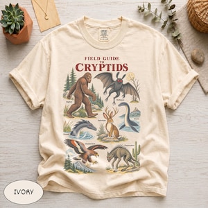 May include: An ivory short-sleeve t-shirt featuring a graphic print titled Field Guide to Cryptids. The design displays illustrations of Bigfoot, Mothman, Jackalope, Nessie, Champ, Thunderbird, and Chupacabra with their names printed below each creature.
