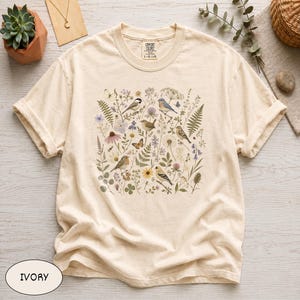 May include: An ivory short-sleeve t-shirt featuring a central graphic print of various birds, butterflies, and wildflowers in muted tones. The shirt has a crew neckline and a Comfort Colors brand label printed on the inside of the collar.