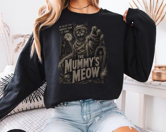 Puede incluir: Una sudadera negra de manga larga con un estampado gráfico de un gato envuelto en vendas como una momia, extendiendo sus garras. El texto dice: AN ANCIENT EVIL AWAKENS FROM ITS TOMB! THE MUMMY'S MEOW, con una calavera y un ataúd debajo.