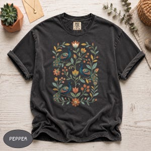 May include: A charcoal gray short-sleeve t-shirt featuring a central graphic print of three peacocks surrounded by stylized floral vines, leaves, and berries in shades of blue, orange, yellow, and green. The shirt has a crew neckline and a label.