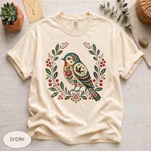 May include: An ivory t-shirt featuring a central graphic of a blue bird adorned with red roses, stars, and intricate patterns, framed by a wreath of green leaves and red berries. The shirt has a crew neckline and short sleeves with rolled cuffs.
