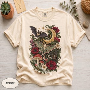 May include: An ivory short-sleeve t-shirt featuring a central graphic print of a moth, a crescent moon, a bat, mushrooms, a spider web, and red flowers. The shirt has a crew neckline and a Comfort Colors brand label printed on the inside collar.