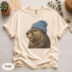 May include: An ivory-coloured short-sleeve t-shirt featuring a graphic illustration of a brown groundhog wearing a blue knit beanie. The shirt has a crew neckline and a Comfort Colors brand label printed on the inside of the collar area.