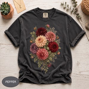 May include: A charcoal gray short-sleeve t-shirt featuring a central graphic of a floral bouquet with dahlias in shades of deep red, light pink, and purple, accented by green leaves and small white filler flowers. The shirt has a crew neckline.