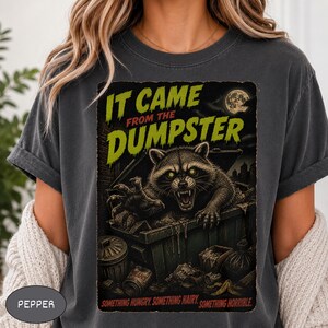 Peut inclure: Un t-shirt gris anthracite avec un motif de raton laveur sortant d'une benne à ordures, aux yeux jaunes brillants et aux crocs apparents. Le texte indique IT CAME FROM THE DUMPSTER et SOMETHING HUNGRY. SOMETHING HAIRY. SOMETHING HORRIBLE. Pleine lune.
