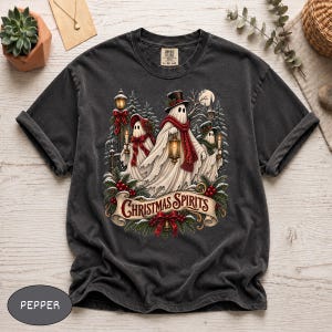 Puede incluir: Una camiseta gris carbón con un gráfico de tres fantasmas con bufandas y sombreros, sosteniendo linternas entre pinos y acebo. El texto "CHRISTMAS SPIRITS" está impreso en una pancarta roja en la parte inferior de la ilustración.