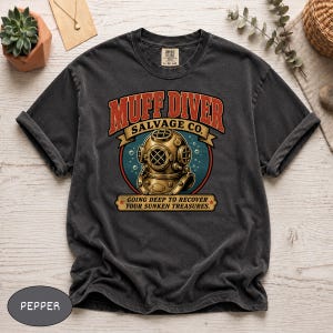May include: Pepper-colored t-shirt with a vintage-style graphic. The graphic features the text "MUFF DIVER SALVAGE CO." above an illustration of a diving helmet. Below the helmet, the text reads "GOING DEEP TO RECOVER YOUR SUNKEN TREASURES."