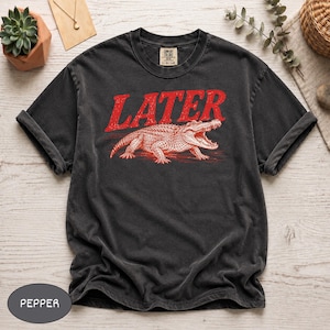 May include: A charcoal grey short-sleeve t-shirt featuring a red graphic of an alligator with its mouth open. Above the alligator, the word LATER is printed in a distressed red font. The shirt has a crew neckline and a vintage-washed appearance.