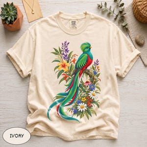 May include: An ivory short-sleeve t-shirt featuring a graphic print of a resplendent quetzal bird with vibrant green, red, and blue plumage perched on a branch surrounded by a colorful arrangement of various tropical flowers and green foliage.