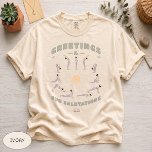 May include: Ivory-colored t-shirt with the words "GREETINGS & SUN SALUTATIONS" in a circular design. The design includes illustrations of yoga poses around a sun graphic. The shirt is a Comfort Colors brand, size L-G/LG.