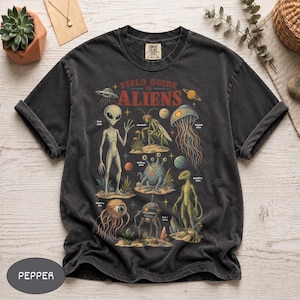May include: A charcoal gray t-shirt featuring a graphic print titled Field Guide to Aliens. The design displays various extraterrestrial creatures including a gray alien, insectoid, jellyfish alien, floating eye, robot, and reptilian alien with text labels.