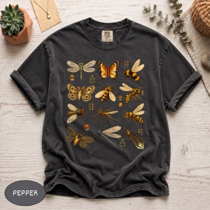 May include: A charcoal gray t-shirt featuring a graphic print of various insects including butterflies, bees, dragonflies, and wasps arranged in a grid pattern. The shirt has short sleeves, a crew neckline, and a label reading Comfort Colors.