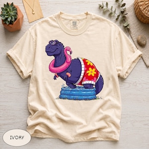 May include: Ivory-colored t-shirt featuring a cartoon dinosaur wearing sunglasses, a pink flamingo float, and a red and yellow floral shirt. The dinosaur is in a blue kiddie pool. The shirt has a Comfort Colors tag.