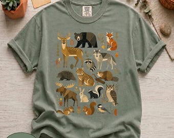 May include: A sage green short-sleeve t-shirt featuring a graphic print of various North American forest animals including a deer, bear, fox, wolf, raccoon, lynx, beaver, porcupine, moose, rabbit, skunk, owl, chipmunk, squirrel, and a small bird.