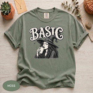 May include: A moss green t-shirt featuring a black and white graphic of a witch wearing a pointed hat and holding a coffee cup. The text "BASIC" is printed in a large, stylized font above the illustration. The shirt has a crew neckline.